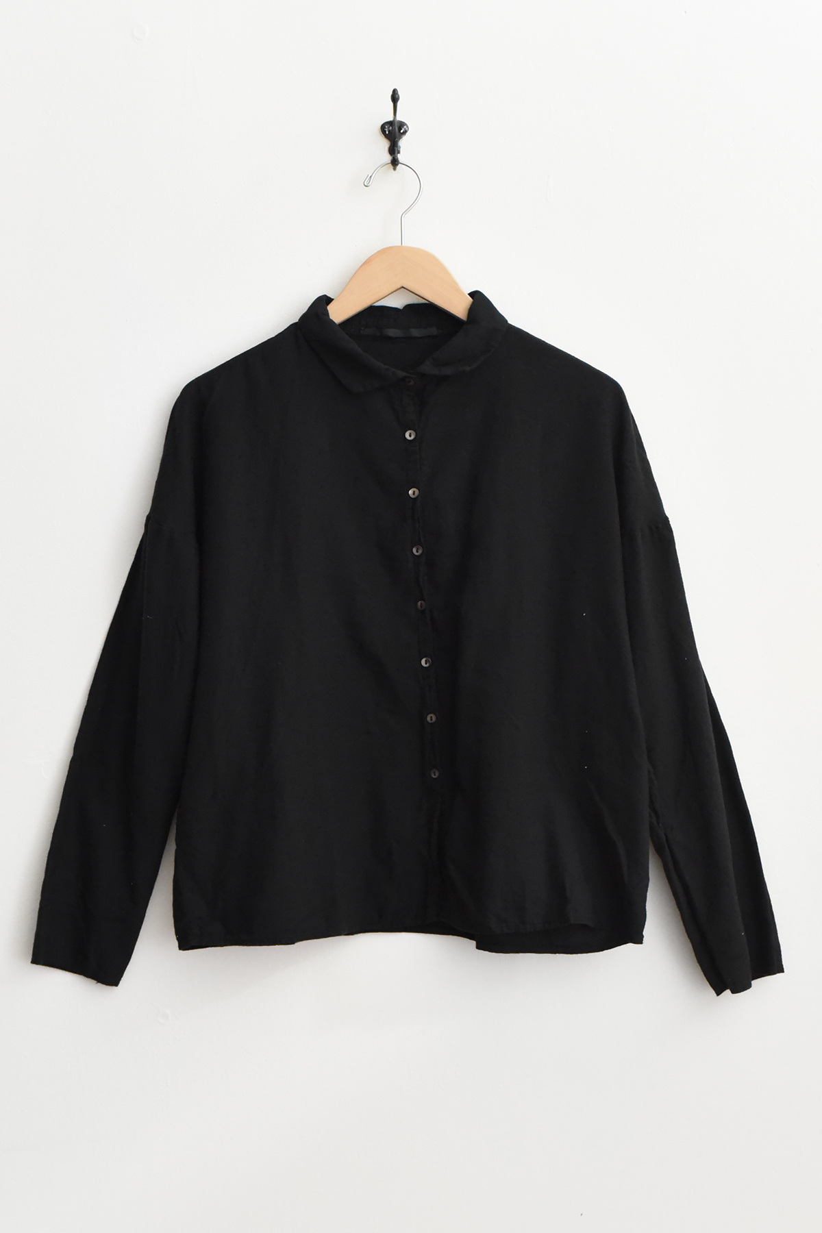 The Album di Famiglia 12173 Short Collar Shirt LWC in black, featuring long sleeves and a button-up design, hangs on a wooden hanger against a white wall. アルバムディファミリア　コットンシャツ