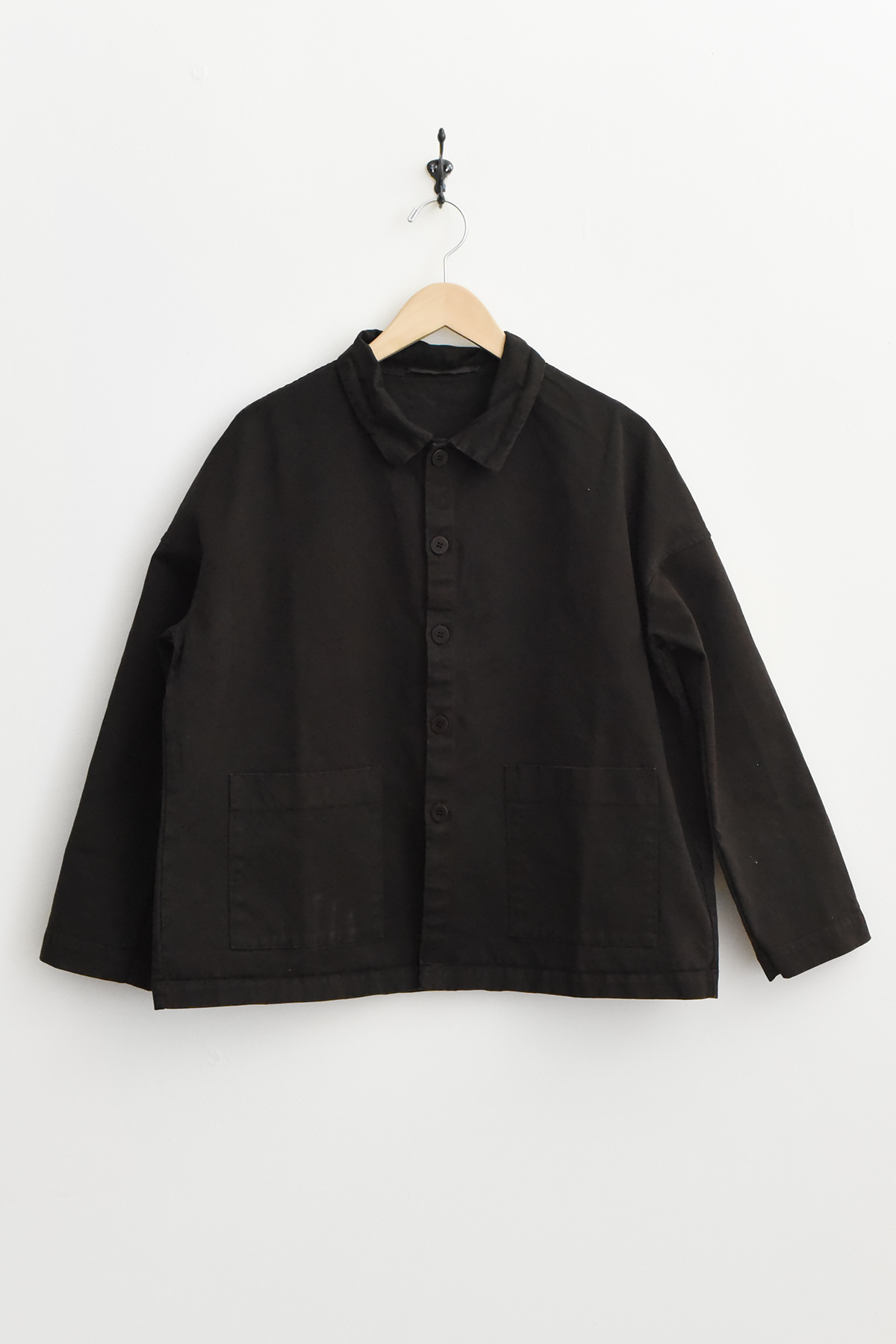 The Album di Famiglia 4286 Overshirt WCC in brown, featuring a button-up front, collared neckline, and two front pockets, hangs on a wooden hanger against a white wall. シャツジャケット