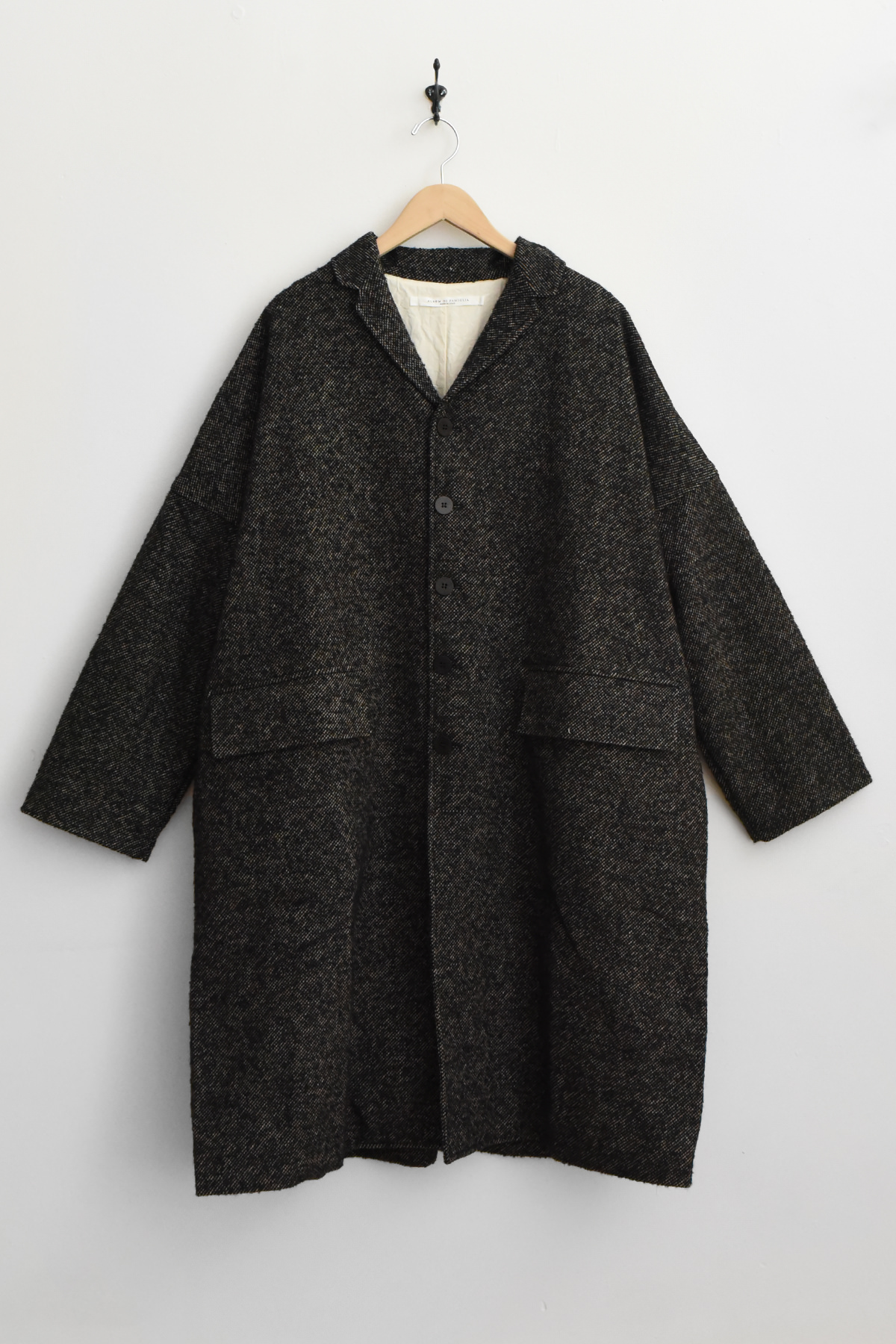The Album di Famiglia U4558 Loose Coat WN in black, featuring a long, relaxed silhouette with a collar and two large front pockets, is displayed on a wooden hanger against a plain white wall.