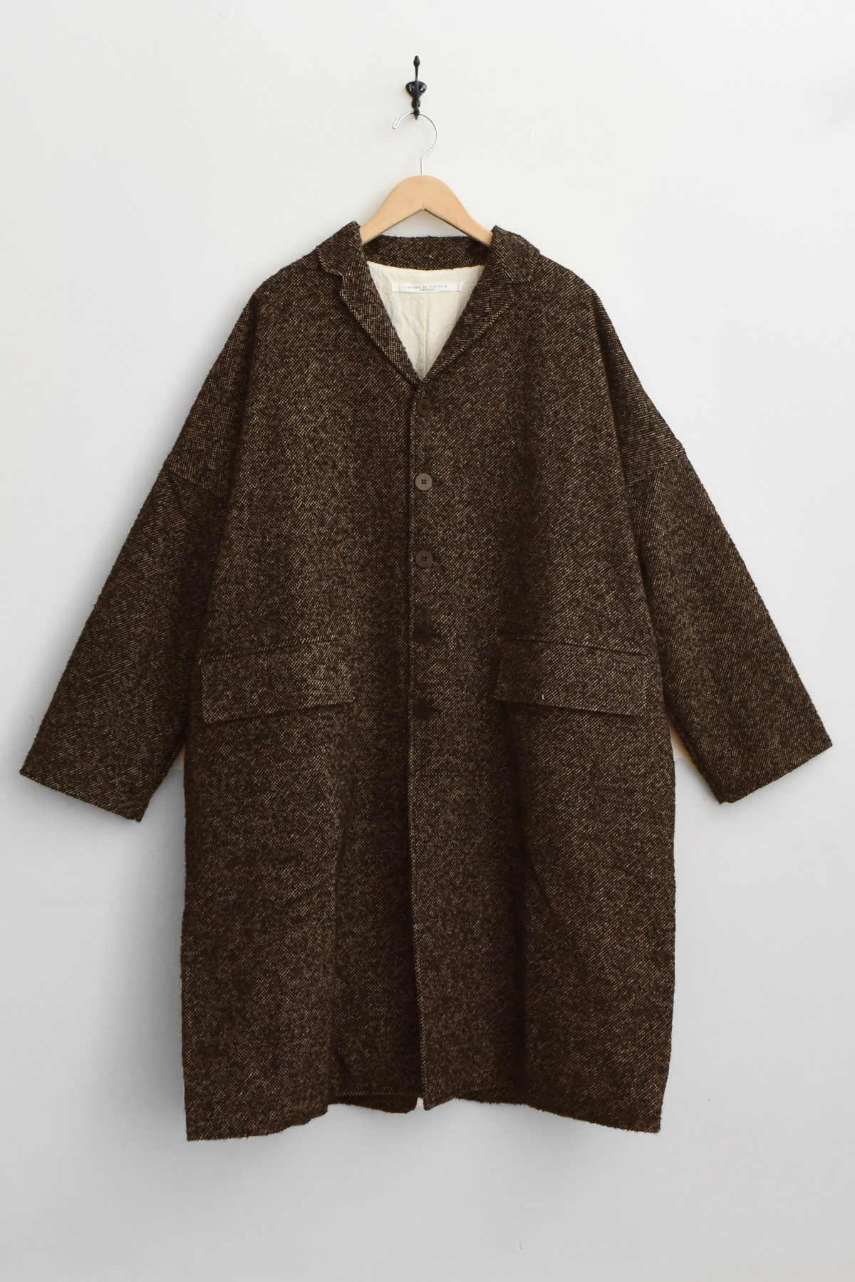 The Album di Famiglia U4558 Loose Coat WN in black, featuring a long, relaxed silhouette with a collar and two large front pockets, is displayed on a wooden hanger against a plain white wall.アルバムディファミリア コート