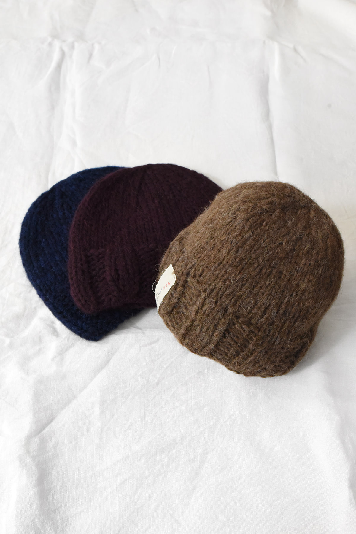 Three Album di Famiglia 9560 Hand Knitted Hats—in brown, navy, and wine—are arranged on a white fabric surface.